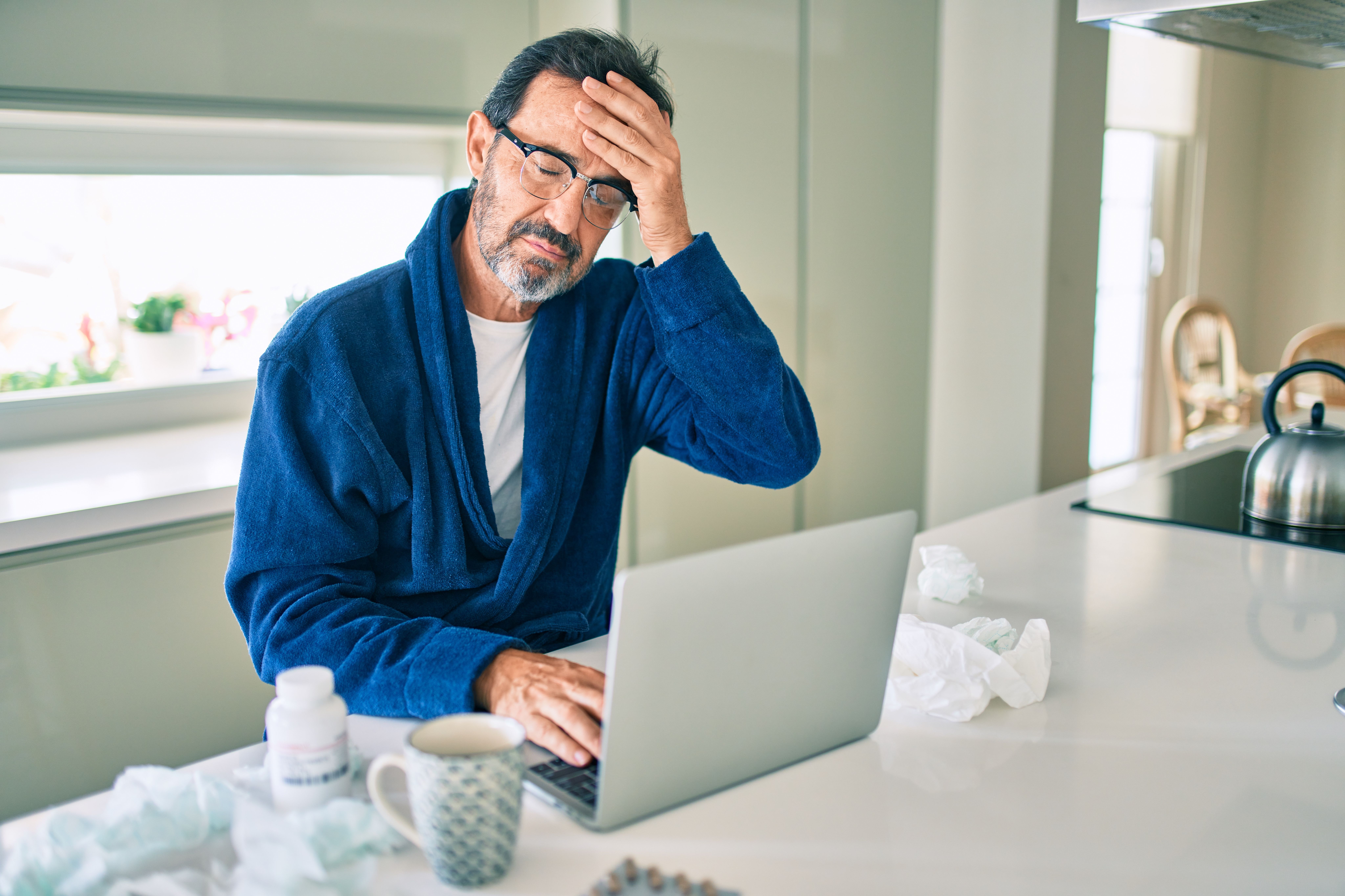 A man grabbing his forehead while trying to work on his laptop at home in his kitchen, struggling with his Cold Agglutinin Disease symptoms.