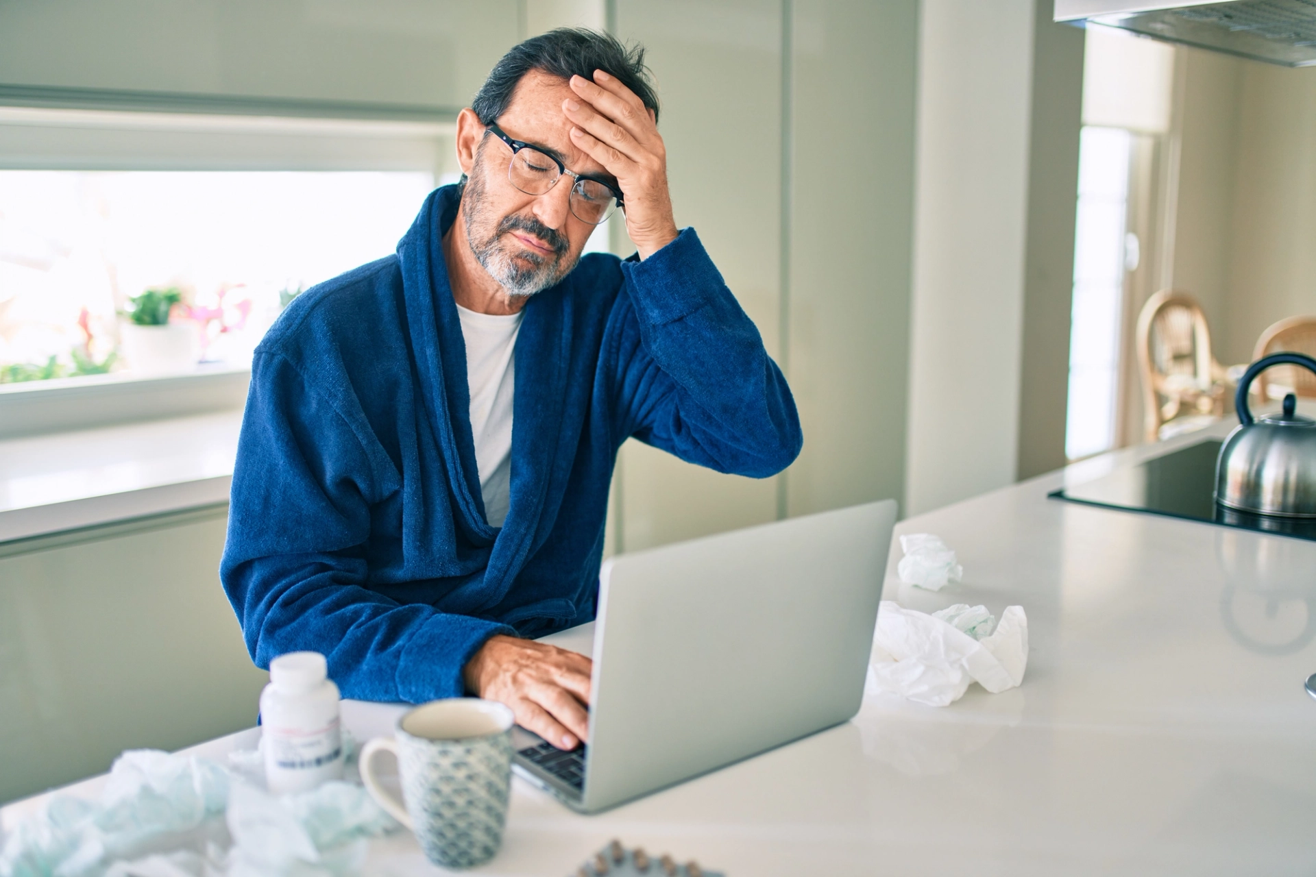 A man grabbing his forehead while trying to work on his laptop at home in his kitchen, struggling with his Cold Agglutinin Disease symptoms.