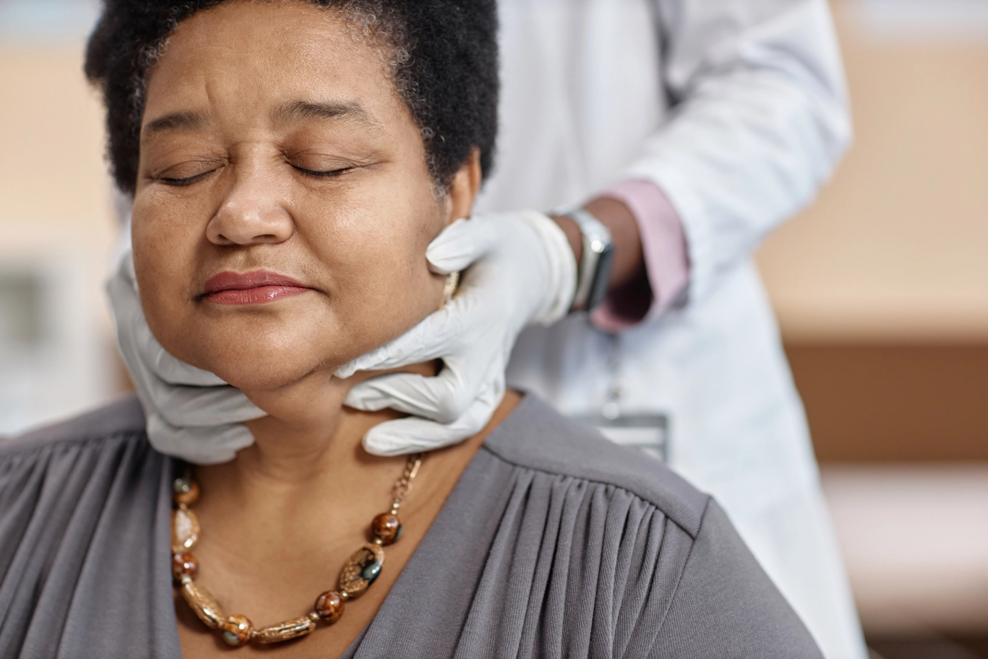 doctor evaluating a female patient's thyroid by feeling around her neck. She is closing her eyes, wearing a beaded necklace, and gray shirt. The doctor is partially out of frame, wearing gloves, an apple watch and feeling around her neck.