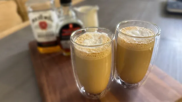Two glasses of maple bourbon latte with foam tops, with liquor bottles blurred in the background.