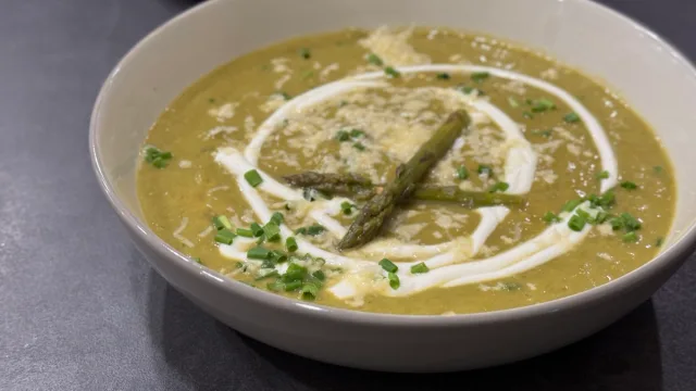 Cream of asparagus soup in a white bowl, garnished with sour cream drizzle, fresh asparagus spears, and chopped chives.
