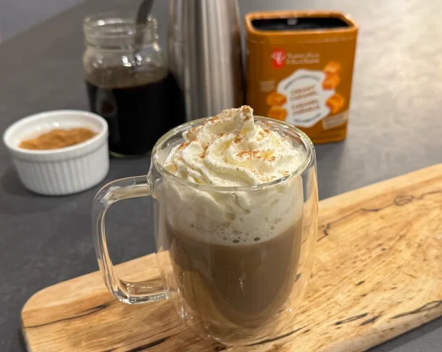 Gingerbread latte in a glass mug topped with whipped cream and spice, surrounded by coffee ingredients and supplies.