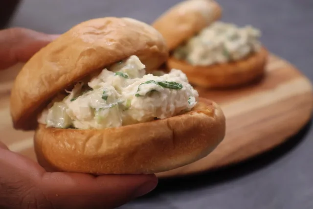 Hand holding a toasted bun filled with creamy crab salad and fresh chives, with more sandwiches on a wooden board.