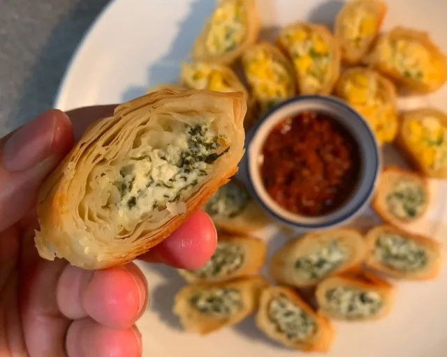 Hand holding a halved spinach cream cheese samosa revealing creamy green filling, with more samosas and red dipping sauce blurred in background.