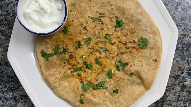 Golden paneer paratha garnished with fresh cilantro, served with a bowl of white yogurt dip on the side.