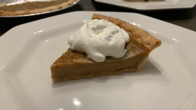 Slice of deep dish pumpkin pie with flaky crust topped with whipped cream on a white plate.