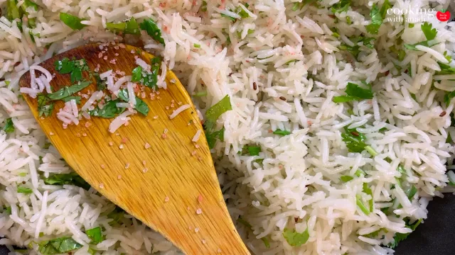 Fluffy jeera rice garnished with fresh cilantro and ginger, stirred with a wooden spoon.