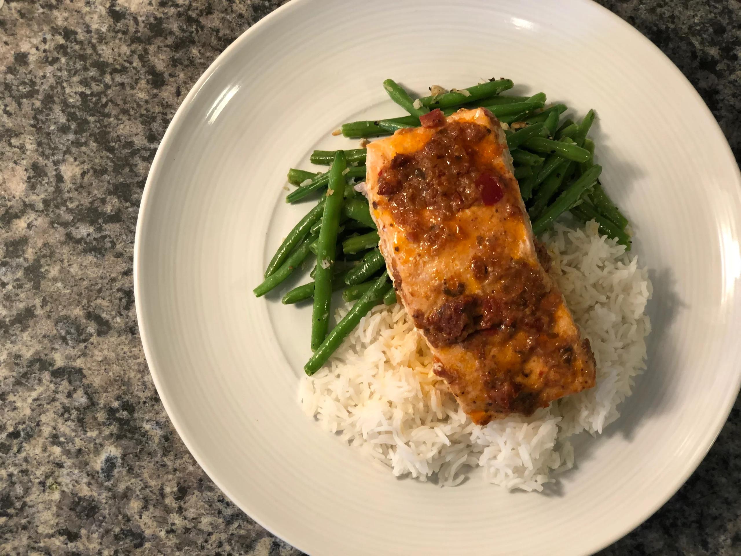 Red pepper pesto salmon fillet with fresh green beans and white rice on a white plate.