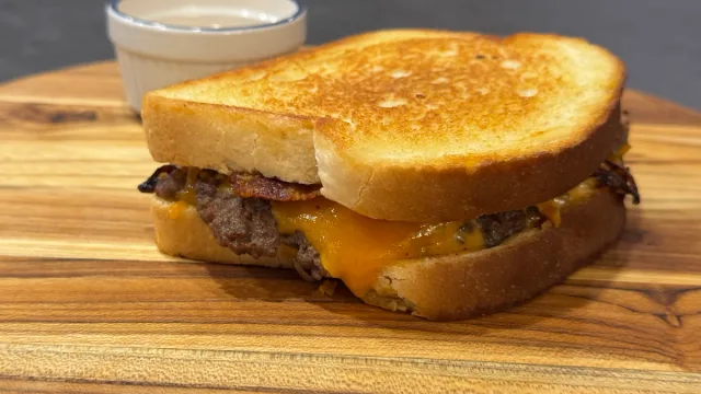Smash burger patty melt with melted cheddar cheese and bacon between toasted bread on a wooden board.