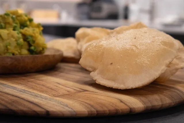 Golden-brown puffy poori bread on wooden board with green chutney in small bowl.