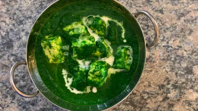 Palak paneer in a green pot with creamy white paneer cheese cubes in spinach sauce.