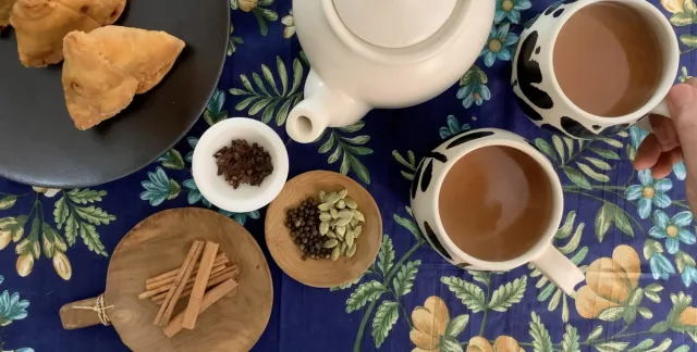 Overhead view of masala chai ingredients and cups: spiced tea with milk, cinnamon sticks, cardamom, cloves, ginger, and fried pastries.