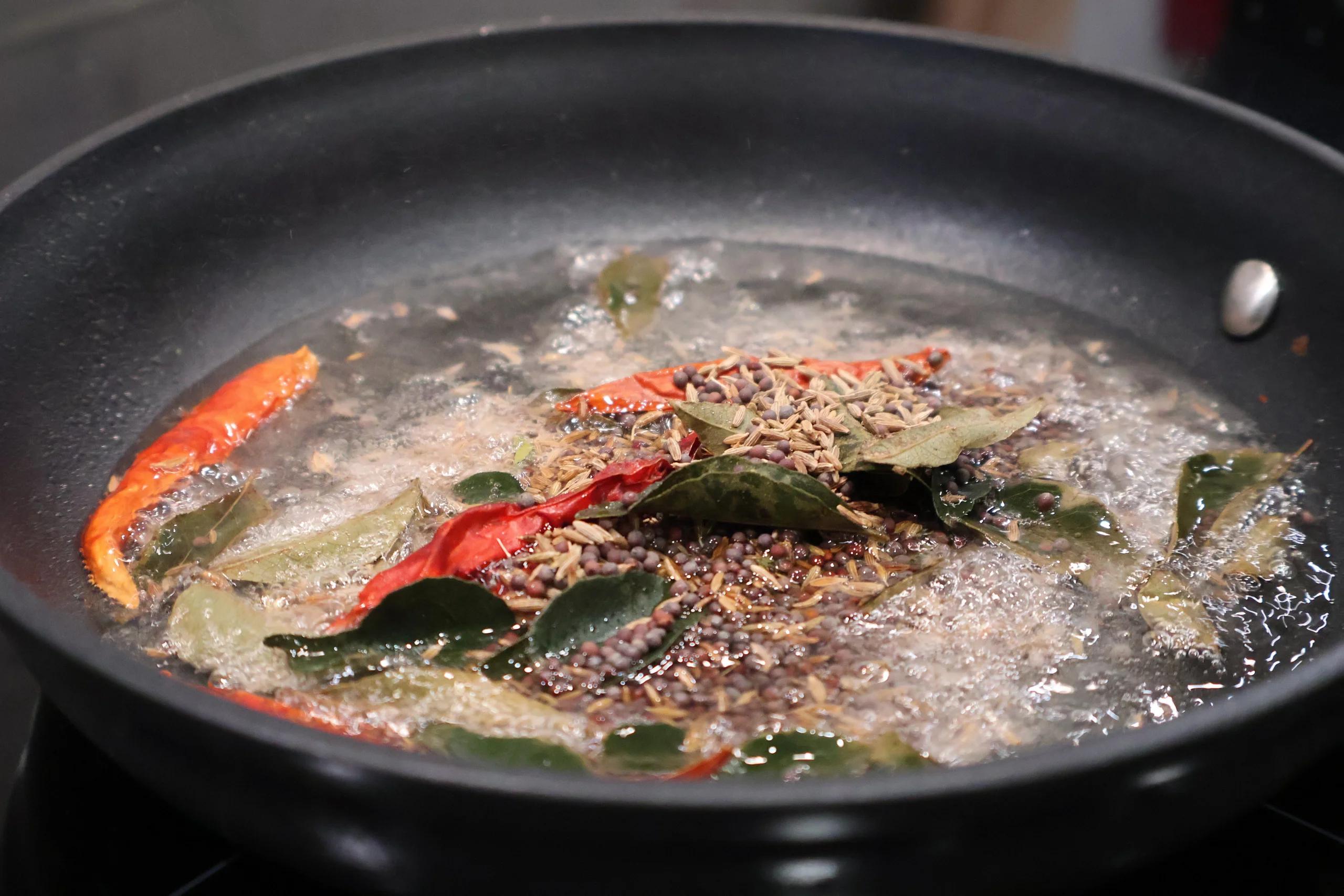 Cumin seeds and mustard seeds blooming in hot oil with red chilies and curry leaves in a dark pan.