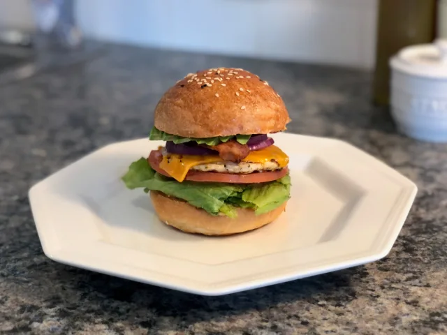 Grilled chicken burger with sesame seed bun, melted cheese, tomato, lettuce, and red onion on a white plate.
