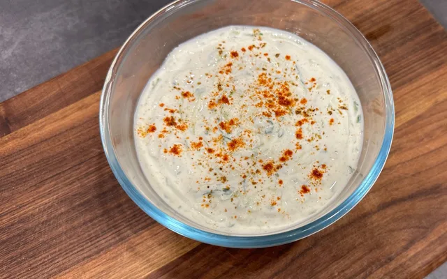 Cucumber raita in a glass bowl topped with paprika and herbs on a wooden surface.