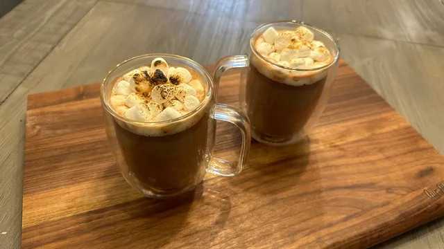 Two mugs of s'mores hot chocolate topped with whipped cream and cocoa powder on a wooden board.