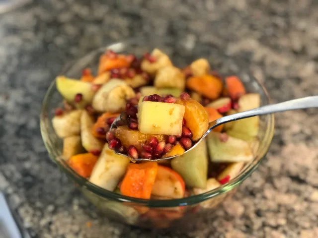 Spoon lifting mixed fruit chaat with diced apples, oranges, and pomegranate seeds from a glass bowl.