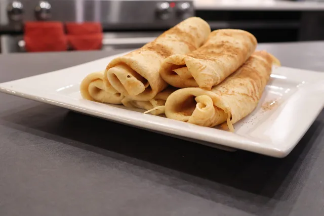 Rolled pumpkin crepes stacked on a white plate in a kitchen setting, dusted with cinnamon.