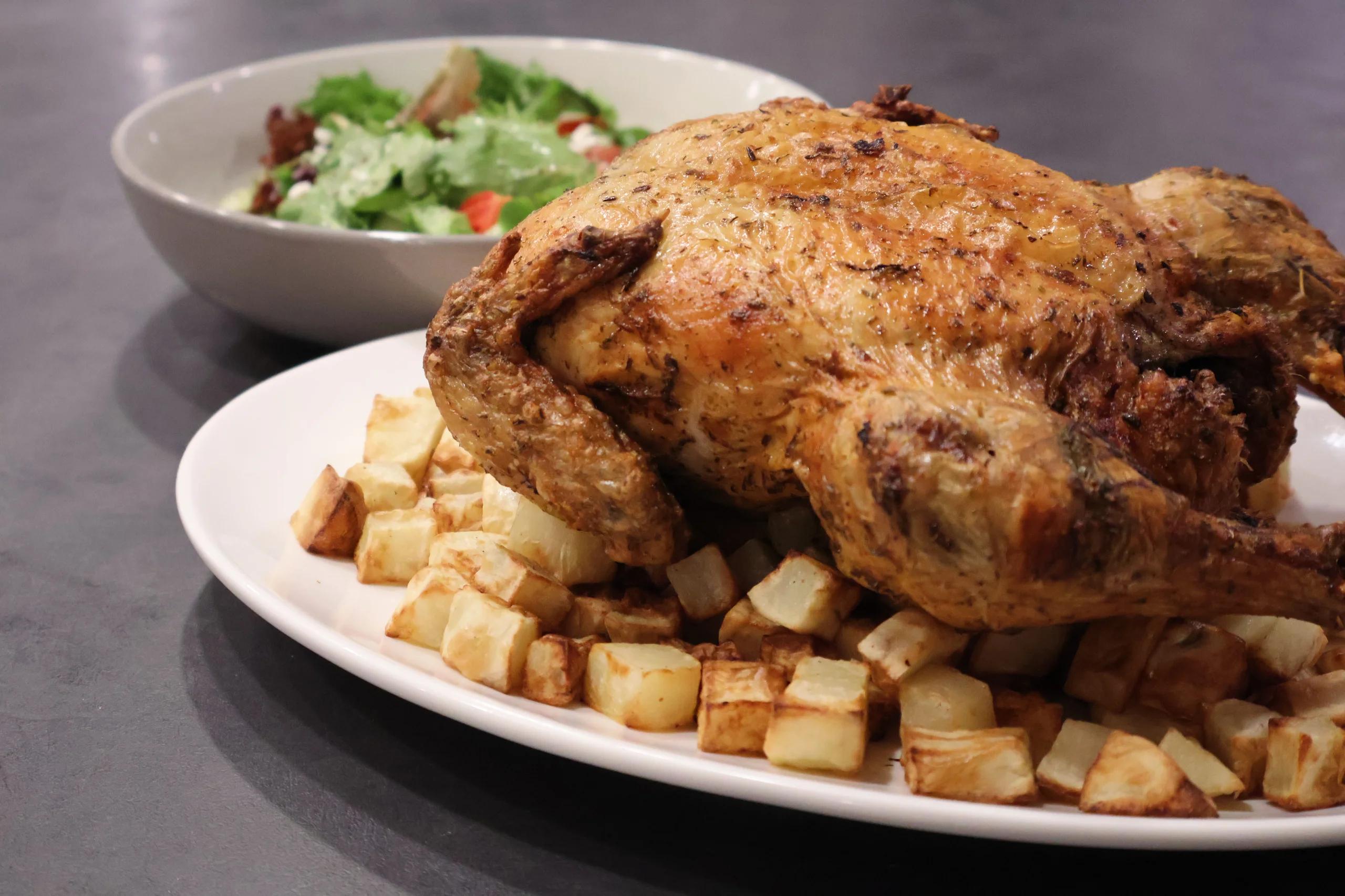 Whole roast chicken with golden, herb-flecked skin on a white platter, ringed with crisp diced potatoes, a green side salad in a bowl behind it.