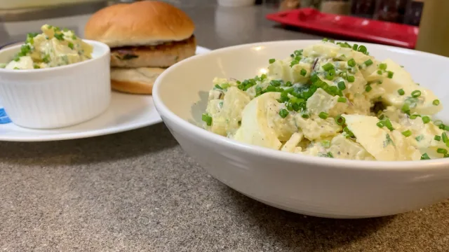 Creamy potato salad with green onions and peas in a white bowl, served with coleslaw and a burger.
