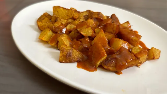 Crispy golden-brown patatas bravas served on a white plate with reddish sauce coating the fried potato cubes.
