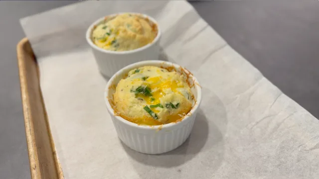 Two baked cheesy egg soufflés in white ramekins, topped with melted cheese and fresh chives.
