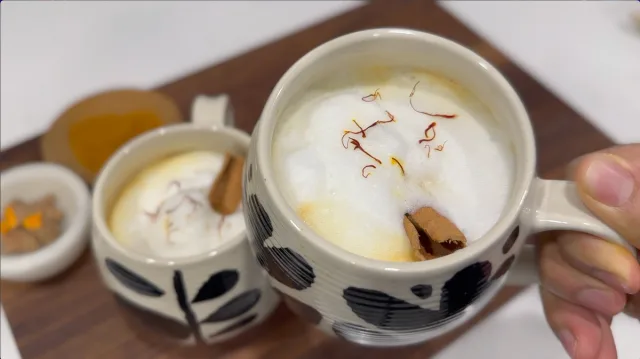 Two ceramic cups of creamy turmeric chai latte topped with saffron strands and cinnamon sticks on a wooden board.