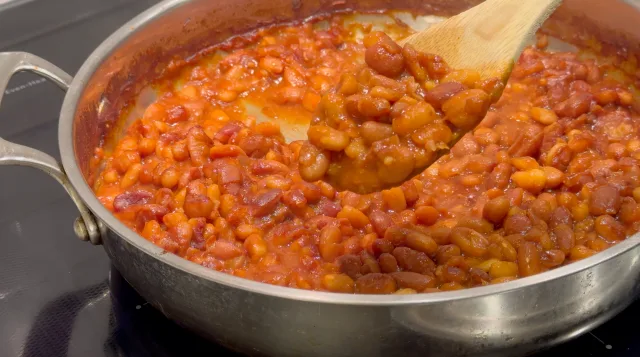 Homemade baked beans in a pot with a wooden spoon, showing beans in tomato sauce.