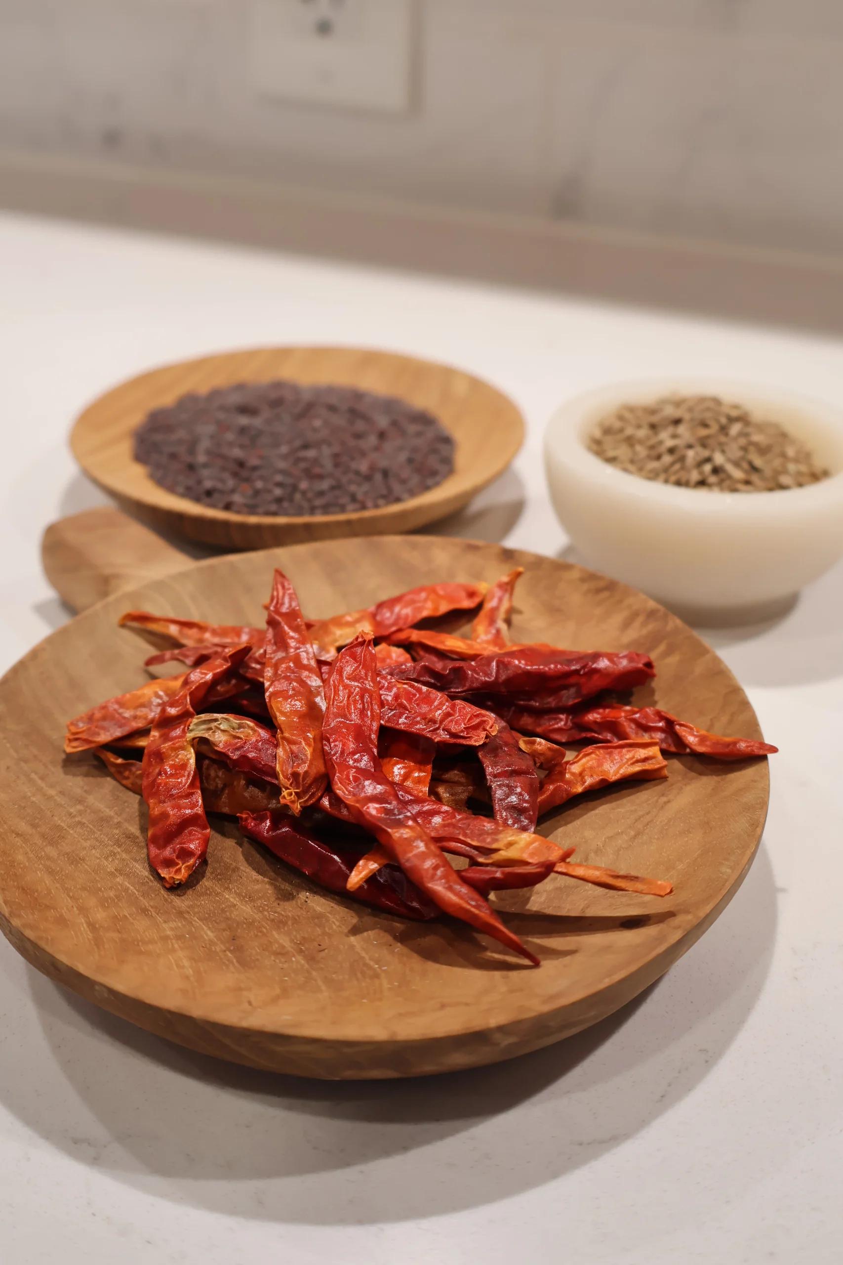 Dried red chili peppers on a wooden plate with bowls of black lentils and cumin seeds in the background.