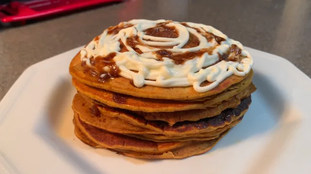 Stack of three pumpkin spice pancakes topped with cinnamon swirl and cream cheese frosting on a white plate.