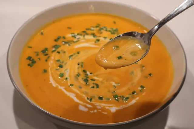Creamy carrot ginger soup with coconut milk garnished with fresh chives, spoon held above bowl.
