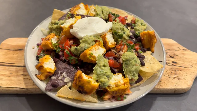 Paneer pakora nachos topped with guacamole, sour cream, pico de gallo, and black beans on a white plate.