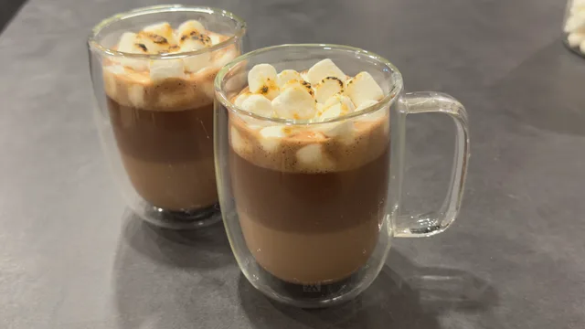 Two glasses of iced chocolate drink topped with whipped cream and chocolate drizzle on gray surface.