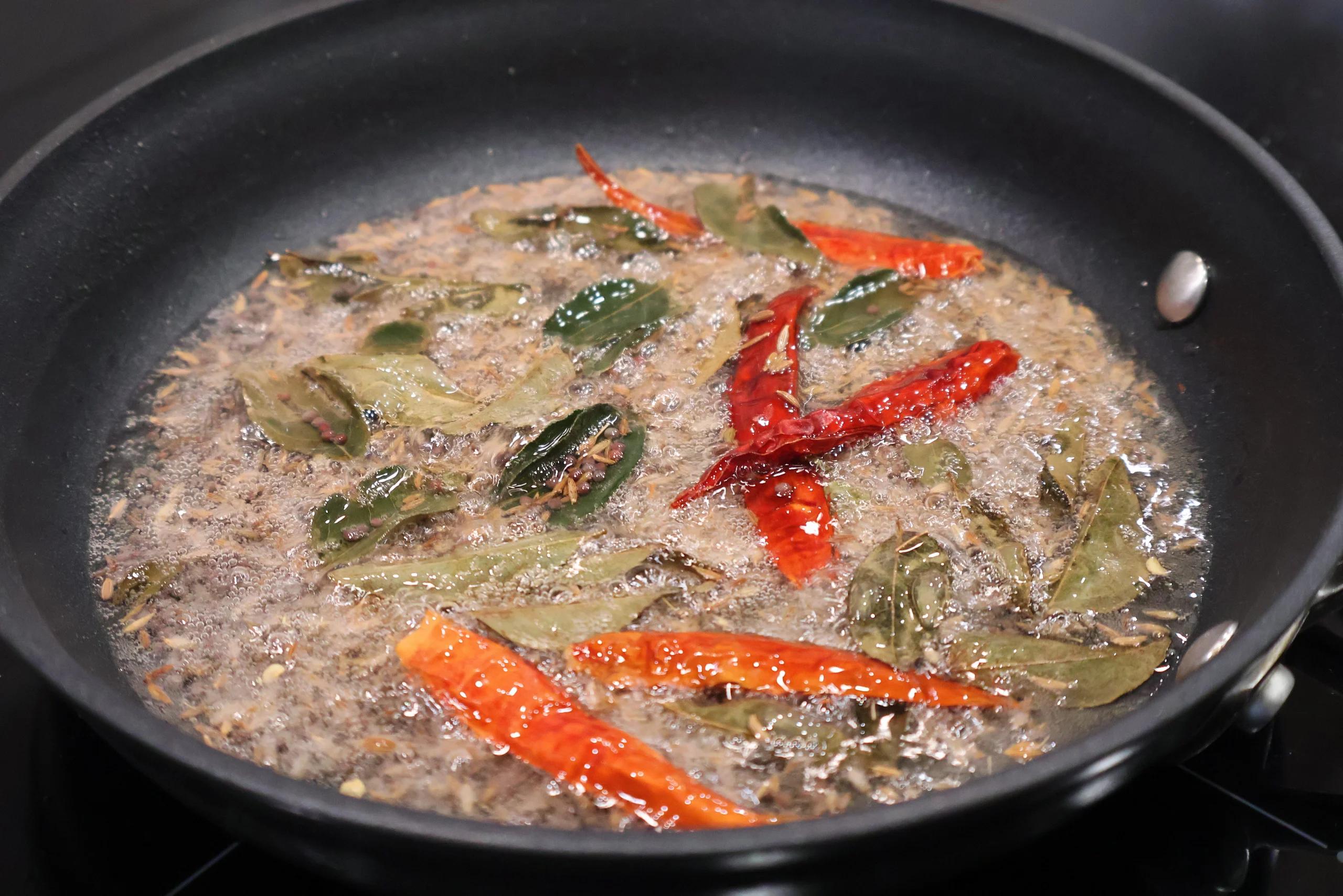 Bloomed spices, sliced red chilies, and green chilies sizzling in oil in a black pan during the aromatic cooking stage.