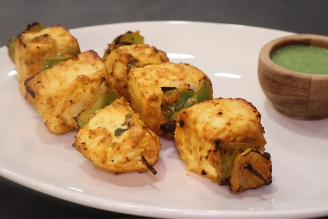 Golden-brown air fryer paneer tikka skewers served on a white plate with green chutney in a wooden bowl.