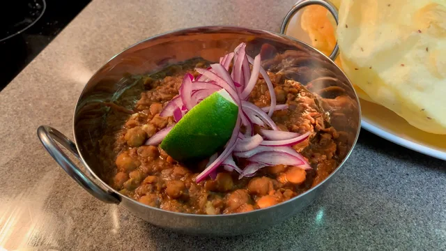 Delhi style chole curry in a steel bowl, garnished with sliced red onions and lime wedge, served with bread.