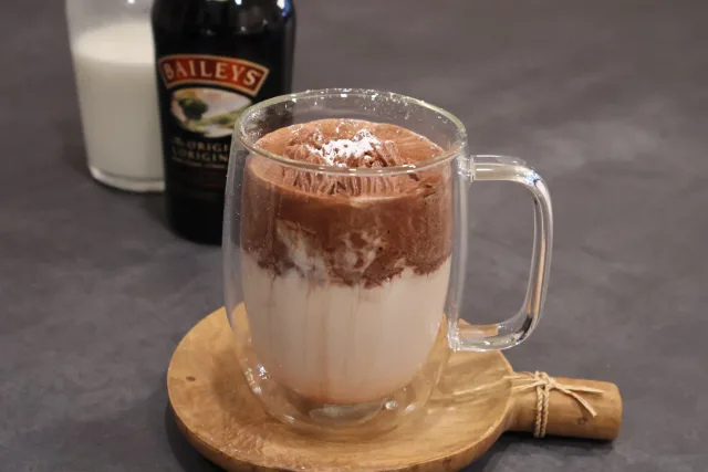 Rich whipped Baileys hot chocolate in a glass mug with cream and cocoa powder on top, beside a bottle of Baileys.