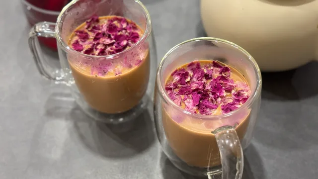Two glass mugs of Gulab Masala Chai with creamy tan liquid topped with dried rose petals and a white pitcher in background.