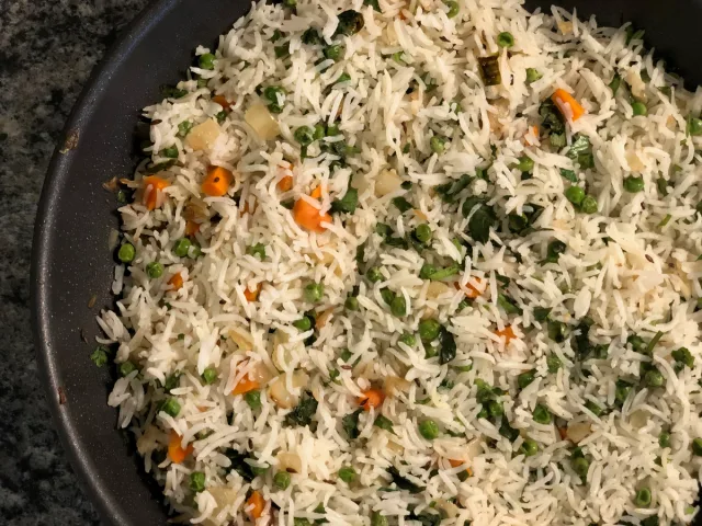 Vegetable rice pulao with white rice, green peas, diced carrots, and onions mixed in a dark skillet.