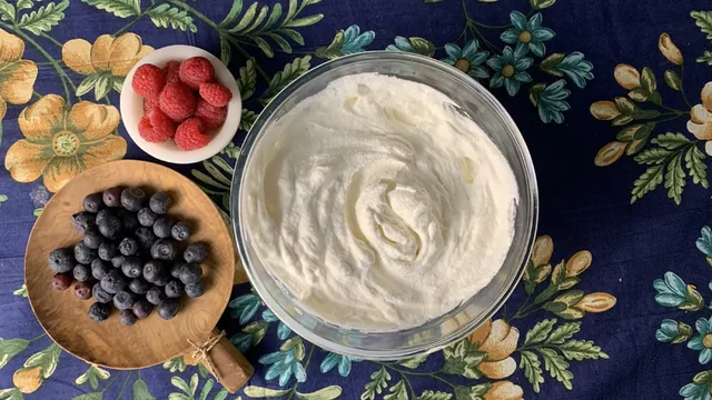 Homemade whipped cream in a glass bowl with fresh raspberries and blueberries on a floral tablecloth.