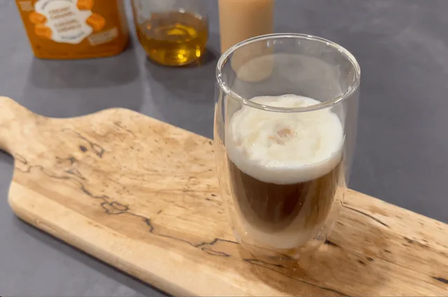 Caramel macchiato in a glass with espresso and milk foam on a wooden board with bottles in background.