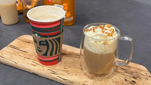 Homemade caramel brulee latte in a glass mug next to an empty Starbucks cup on a wooden board.