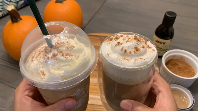 Two pumpkin spice frappuccinos with whipped cream and cinnamon dust, held in hands beside pumpkins and ingredients.
