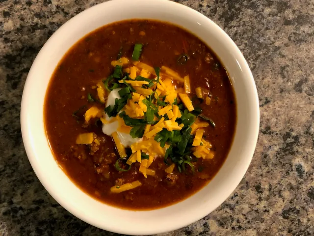 Bowl of beef chili topped with shredded cheddar cheese and fresh green onions on granite countertop.