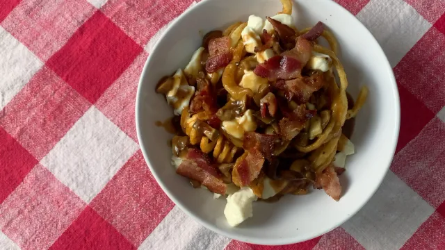 Poutine with crispy fries, melted cheese curds, bacon pieces, and mushrooms in a white bowl on a red gingham cloth.