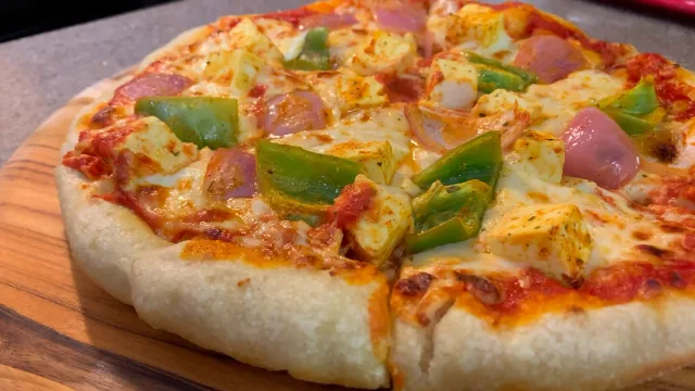 Indian Fusion Paneer Tikka Pan Pizza with melted cheese, paneer cubes, and green peppers on wooden board.