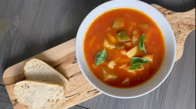 Bowl of vegan minestrone soup with vegetables and broth, garnished with basil, served with crusty bread.