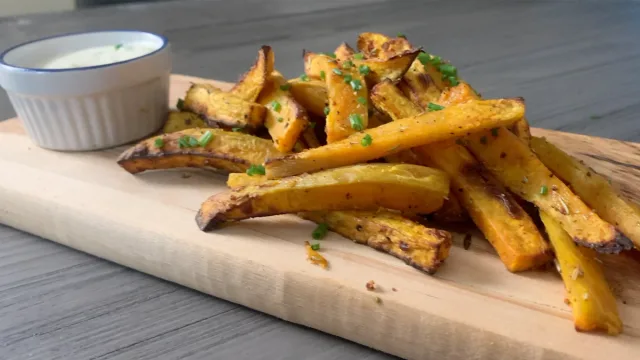 Crispy air fryer sweet potato fries on a wooden board with fresh herbs and a white dipping sauce.