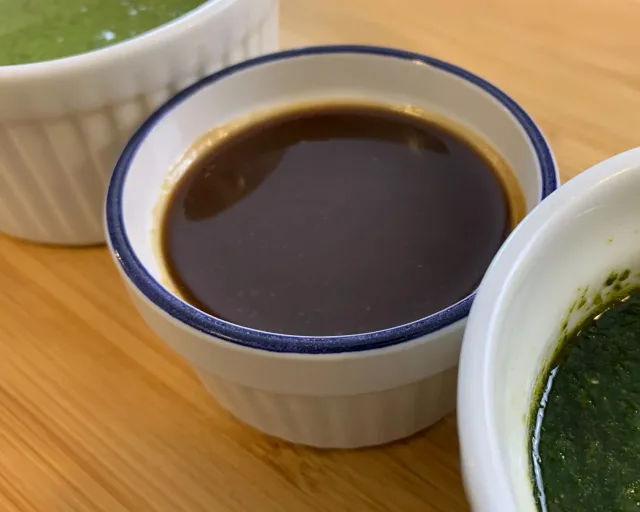 Three bowls of chutneys on a wooden surface: dark tamarind chutney, green cilantro chutney, and light-colored chutney.