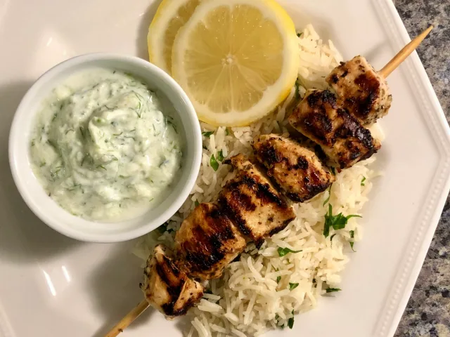 Grilled Greek souvlaki chicken skewers on rice with tzatziki sauce and lemon slices.
